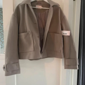 Beige jacka med knappar - Säljer en snygg beige kappa med knappar framtill. Den har en klassisk krage och stora fickor på framsidan. Ärmarna är långa och har dekorativa knappar vid ärmsluten. Perfekt för höst och vår!