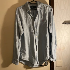 Ljusblå skjorta från Ralph Lauren - Säljer en ljusblå skjorta från Ralph Lauren. Skjortan är långärmad och har en liten röd broderad logga på bröstet. Den är gjord av bomull och har knappar hela vägen ner. Observera att det finns en reva på ena ärmen. Perfekt för en avslappnad stil! Storlek M men skulle säga att den passar mer som L. Hör av dug om du har frågor!