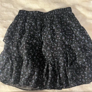 Svart blommig kjol med volanger - Säljer denna superfin svart kjol med små blommor i olika färger. Kjolen har volanger. Perfekt för både vardag och fest! Den är i ett lätt och luftigt material. Säljer för 100kr men pris kan alltid diskuteras. Vid frågor hör gärna av dig!❣️