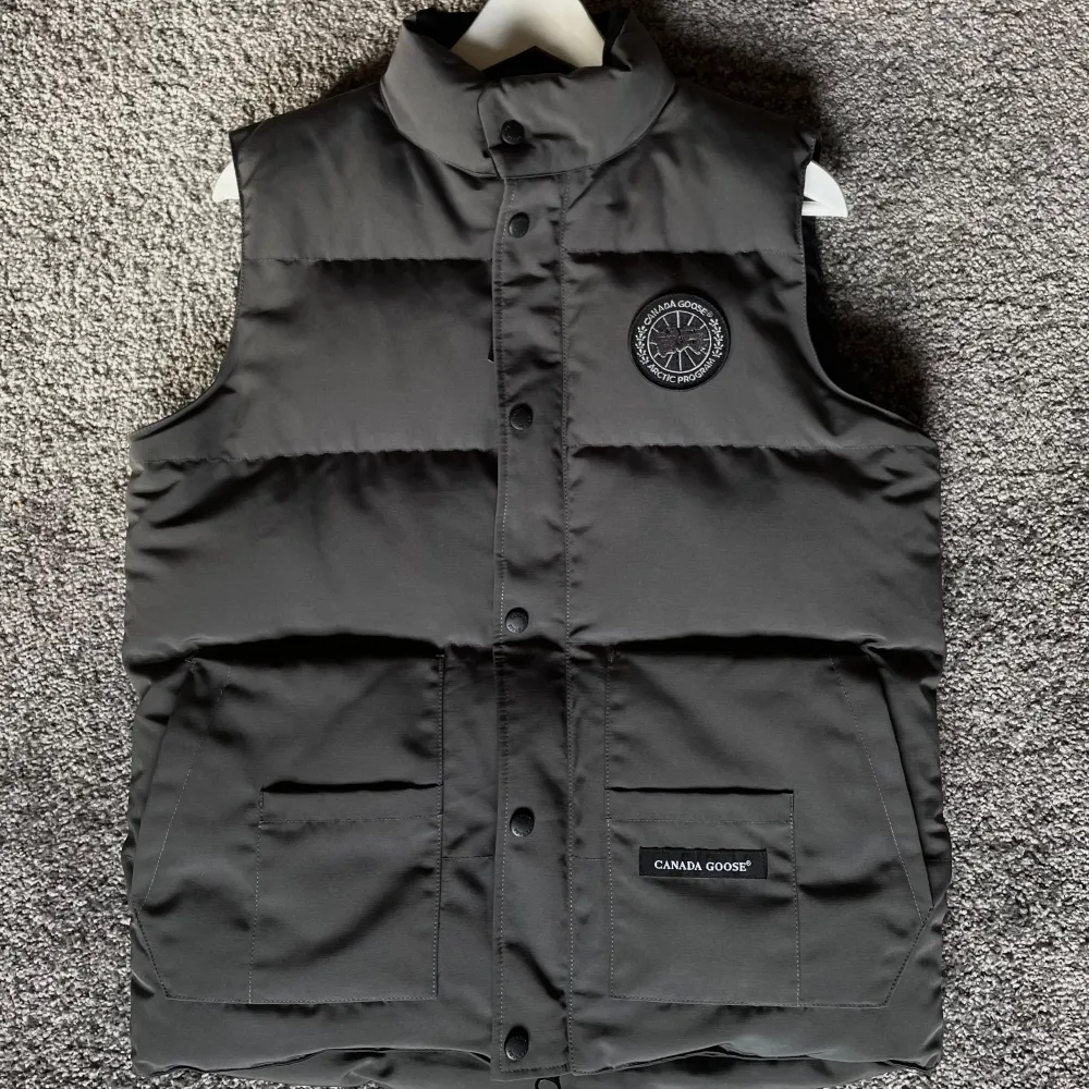 Säljer en snygg och varm svart dunväst från Canada Goose. Västen har en quiltad design med både dragkedja och knappar framtill. Perfekt för kyliga höst- och vinterdagar. Den har också praktiska fickor och en stilren logga på bröstet.. Jackor.