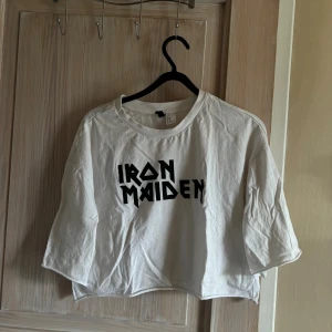 Vit Iron Maiden t-shirt från Divided - Säljer en cool vit t-shirt från Divided med Iron Maiden-tryck på framsidan. Den är croppad och har korta ärmar, perfekt för en avslappnad stil. T-shirten är i bomull och känns skön mot huden. Perfekt för sommarens festivaler eller bara hänga med kompisar!