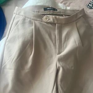 Beige kostymbyxor från Gina Tricot - Säljer ett par stilrena beige kostymbyxor från Gina Tricot. De är högmidjade (men är man mindre än M så kan man ha dem som låg/mid waist) med en knapp och dragkedja framtill. Byxorna har en klassisk rak passform och är perfekta för både vardag och fest. Materialet känns mjukt och bekvämt. 
