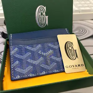 Säljer en snygg och stilren blå plånbok från Goyard. Den har ett klassiskt mönster i blått och vitt, perfekt för att hålla dina kort och kontanter organiserade. Plånboken är i nyskick och kommer i originalförpackning.