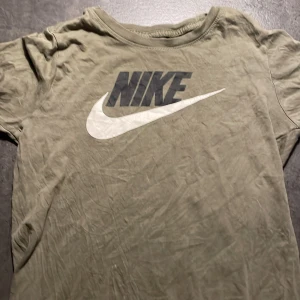 Olivgrön t-shirt från Nike - Säljer en olivgrön t-shirt från Nike med det klassiska logotyptrycket i svart och vitt på framsidan. T-shirten är kortärmad och gjord i ett mjukt bomullsmaterial. Perfekt för en avslappnad stil eller träning. Lite skrynklig men i bra skick!