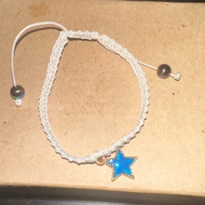 Vitt armband med blå stjärna - Säljer ett supergulligt vitt armband med en blå stjärna som hänge. Armbandet är justerbart med en dragsko och har små pärlor i ändarna. Perfekt för att ge lite extra stil till din outfit!