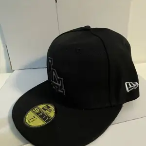 Säljer en snygg svart keps från New Era, modell 59FIFTY. Den har en broderad logga på framsidan och en liten logga på sidan. Kepsen är i bra skick och perfekt för både vardag och sport, 7/1/8 56.8 cm.