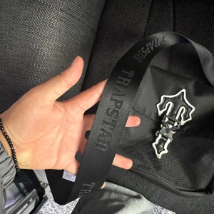 Svart axelväska från Trapstar - Säljer en snygg svart axelväska från Trapstar. Väskan har ett coolt spänne framtill och justerbar axelrem med märkeslogga. Perfekt för att bära dina essentials med stil! 