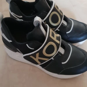 Svarta sneakers med vit sula och logga - Snygga michel kors  stolek 33 svarta sneakers med en vit sula och breda remmar över foten med en stor logga. De har en modern design med vita detaljer som ger en cool kontrast. Perfekta för en casual look eller när du vill sticka ut lite extra.