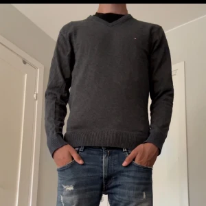 Grå tröja från Tommy Hilfiger - Säljer en stilren grå tröja från Tommy Hilfiger i storlek M. Tröjan är i bomull och kashmir, vilket gör den supermjuk och bekväm. Den har en klassisk v-ringning och långa ärmar, perfekt för både vardag och lite finare tillfällen. Passar perfekt till jeans eller chinos!