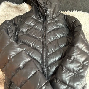 Svart pufferjacka från Hollies - Säljer en snygg svart pufferjacka från Hollies i storlek 36. Jackan är perfekt för kalla dagar med sitt quiltade mönster och dragkedja framtill. Den har en hög krage för extra värme och stil. Passar perfekt för både vardag och festliga tillfällen!