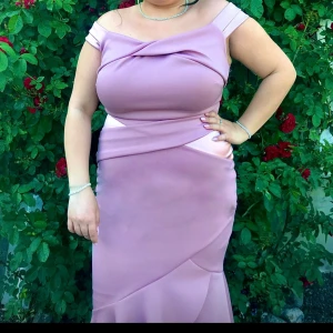 Rosa långklänning för bröllop - Säljer en superfin rosa långklänning från LIPSY. Den har en elegant offshoulder-design med volangdetaljer som ger en snygg touch. Klänningen är perfekt för festliga tillfällen och har en figurnära passform. Materialet känns lyxigt och har en lätt glansig finish. Perfekt för bal eller en speciell kväll! Klänning är använd endast 1 gång för ett bröllop då jag var brudtärna. Sista bilden är en mer tydlig bild på hur modellen ser ut (dock i en annan färg!)