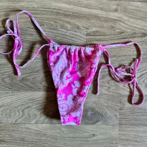 Rosa bikiniunderdel från SHH - Säljer en superfin rosa bikiniunderdel från Shein. Den har ett snyggt paisleymönster i vitt och knytband på sidorna för perfekt passform. Perfekt för sommarens alla badäventyr! 🌸 Aldrig använda! 