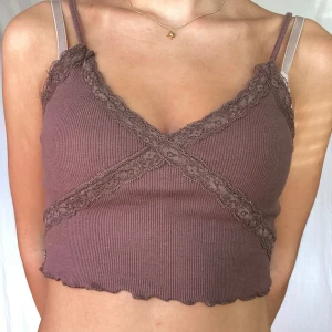 Brun ribbad linne med spetsdetaljer - Säljer en superfin brun ribbad linne med spetsdetaljer. Den har en croppad passform och smala axelband. Perfekt för sommardagar eller som ett lager under en kofta. Spetsen ger en söt touch och den är riktigt bekväm att ha på sig.