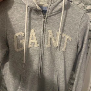 Grå hoodie från GANT - Säljer en snygg och bekväm grå hoodie från GANT. Den har en dragkedja framtill och stora bokstäver i vitt på bröstet. Perfekt för en avslappnad stil och passar till alla tillfällen. Den är i bra skick och redo för en ny ägare! (äkta)