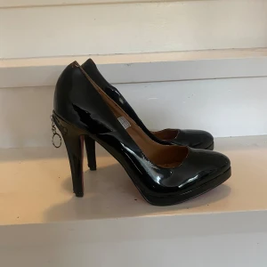 Lackpumps - Oanvända svarta lackpumps med röd sula samt dragkedja detalj på klacken.