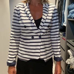 Randig kavaj från Gina Tricot - Säljer en snygg randig kavaj från Gina Tricot i storlek 34 (XS). Kavajen är vit med mörkblå ränder och har en dubbelknäppning framtill med svarta knappar. Den har långa ärmar och är perfekt för både vardag och fest. Passar bra till både jeans och kjol!