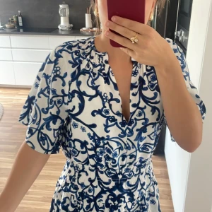 Vit och blå kortklänning med blommigt mönster - Säljer en superfin vit och blå kortklänning med ett blommigt mönster. Klänningen har korta ärmar och en v-ringad hals. Den är perfekt för sommar och vår, och passar bra till både fest och vardag. Materialet känns lätt och bekvämt, och klänningen har en normal passform. Perfekt för att känna sig både snygg och bekväm!