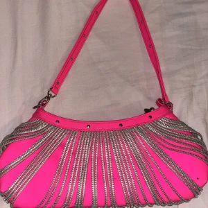 Axelväska med kedjedetaljer - Rosa axelväska med silverfärgade kedjedetaljer. Väskan har en justerbar axelrem och dekorativa nitar längs kanten.