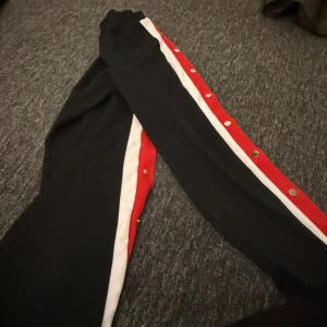 Svarta byxor med röda och vita detaljer från Parisian - Snygga svarta byxor från Parisian med röda och vita ränder längs sidorna. Byxorna har tryckknappar längs benen för en cool look. Perfekta för en sportig stil.