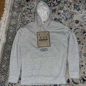 Grå hoodie från Burberry - Snygg grå hoodie från Burberry med klassisk logga på bröstet. Tröjan har en bekväm passform med känguruficka och justerbar huva. Perfekt för en stilren och avslappnad look. Pris kan sänkas vid snabb affär
