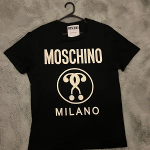 Svart t-shirt från Moschino - Snygg svart t-shirt från Moschino med stort vitt tryck på framsidan. Perfekt för en stilren och modern look. T-shirten har en klassisk passform och är kortärmad. Storlek M men skulle säga den passar S. Trycket har lite små skavanker men som man inte lägger märke till vid användning. 