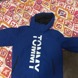 Blå jacka från Tommy Hilfiger - Snygg blå jacka från Tommy Hilfiger med stor logga på framsidan. Jackan har en huva och dragkedja framtill. Perfekt för kyligare dagar.