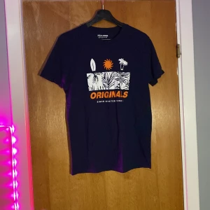 Mörkblå t-shirt från Jack & Jones - Snygg mörkblå t-shirt från Jack & Jones Originals med ett tryck av en surfbräda, sol och palm i vitt och orange. Perfekt för en avslappnad stil.