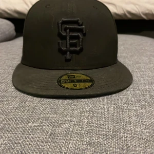 Svart keps från New Era - Säljer en svart keps från New Era med broderad logga framtill och World Series-märke på sidan. Kepsen är i bra skick och gjord av 100% polyester. Perfekt för sportevenemang eller som en stilren accessoar till vardagsoutfiten.