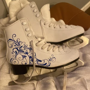 Vita konståkningsskridskor med blått mönster - Säljer ett par vita konståkningsskridskor i mycket bra skick. De har ett snyggt blått, blommigt mönster på sidan och snörning framtill. Perfekta för vinterns isäventyr! Bladen är i rostfritt stål och ger bra grepp på isen.