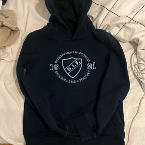 Mörkblå hoodie från Djurgårdens IF - Säljer en mörkblå hoodie från Djurgårdens IF med tryck på bröstet. Tröjan har en klassisk känguruficka och en bekväm huva. Perfekt för alla Djurgården-fans! 💙 jag är storlek 152 i barn och XXS i vuxen och jag skulle säga att denna passar mig men är lite liten så nån som är mindre skulle de passa till 10-12 år
