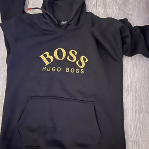 Svart hoodie från Hugo Boss - Säljer en snygg svart hoodie från Hugo Boss med deras logga i guld på bröstet. Använd 1 gång och är i nyskick säljer pga storleken. Storleken är M men passar även som S. 