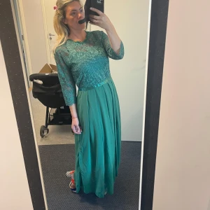 Grön långklänning med spetsdetaljer - Elegant grön långklänning med spetsdetaljer på överdelen och trekvartsärmar. Kjolen är flödande och ger en vacker siluett. Perfekt för speciella tillfällen.Helt ny.