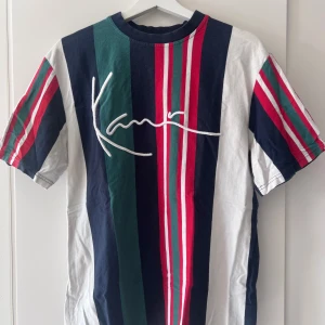 Randig t-shirt från Karl Kani - Snygg randig t-shirt från Karl Kani i färgerna blå, grön, röd och vit. T-shirten har korta ärmar och ett stort tryck med märkesnamnet på framsidan. Perfekt för en avslappnad stil.