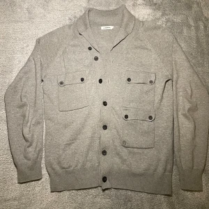 Grå kofta från J.Lindeberg - Snygg grå kofta från J.Lindeberg med knappar och två stora fickor framtill. Perfekt för en stilren look. Storlek M men är lite liten i storlek så kan passa S