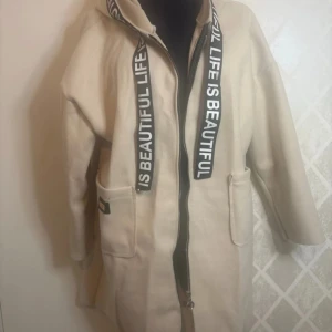 Beige kappa med textdetaljer - Snygg beige kappa med huva och texten 'Life is Beautiful' längs dragkedjan. Kappan har långa ärmar och praktiska fickor framtill. Perfekt för en stilren look.