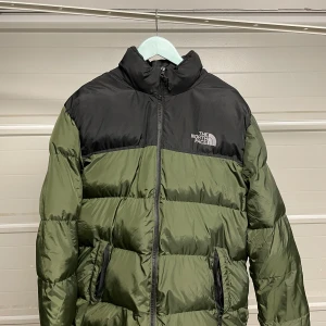 Grön och svart dunjacka från The North Face - Säljer en grön och svart dunjacka från The North Face i mycket bra skick. Jackan har en dragkedja framtill och är perfekt för kalla vinterdagar. Den är långärmad och har en klassisk pufferstil som håller dig varm och bekväm. Perfekt för både vardag och utomhusaktiviteter! Säljes i befintligt skick. Storlek: XL/L