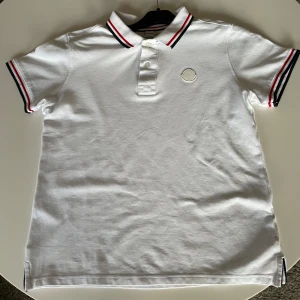 Vit pikétröja från Moncler - Snygg vit pikétröja från Moncler med klassisk krage. Tröjan har korta ärmar där det finns charmiga röd svarta ränder. Perfekt för en stilren look. Hör av er av minsta funderingar😇