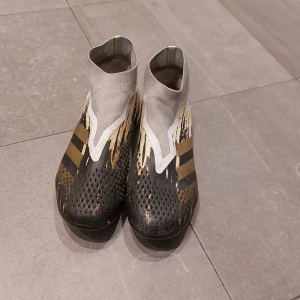 Adidas predator i svart grå och guld. - Säljer ett par coola Adidas predator i grått och svart med guldiga detaljer. Skorna har en strumpliknande passform utan snörning, vilket ger en tight och bekväm känsla på planen. Perfekta för fotbollsspelare som vill ha stil och funktion i ett! Nypris 2000kr