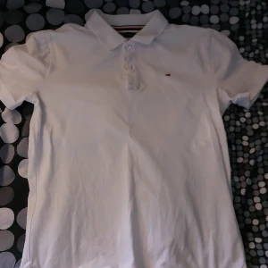Vit pikétröja från Tommy Hilfiger - Säljer en klassisk vit pikétröja från Tommy Hilfiger i bra skick. Den har korta ärmar och en stilren krage med knappar. Perfekt för både vardag och festliga tillfällen. Tröjan har en liten logga på bröstet och snygga detaljer vid sidoslitsarna. Skick 7,5/10 Den har ett pyte litet hål i armhålan. Det går dock att fixa väldigt enkelt. Nypris 600kr
