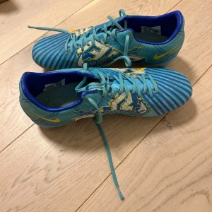 Blå fotbollsskor från Nike - Snygga blå fotbollsskor från Nike med ett randigt mönster och gula detaljer. Skorna har snörning och en sportig design som passar perfekt för fotbollsplanen.