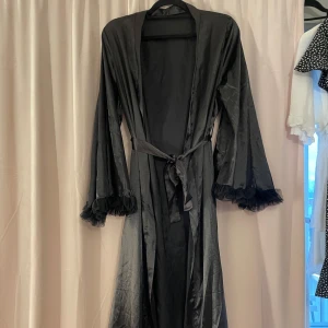 Svart morgonrock  - Aldrig använd! Elegant svart morgonrock i satin med långa ärmar och volangdetaljer vid ärmslut och där nere. Morgonrocken har ett knytband i midjan för en smickrande passform. Hör av dig för mer information💕