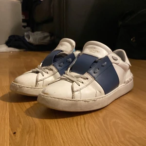 Valentino sneakers med blå detalj - Snygga Valentino sneakers i vitt med en bred blå rem över snörningen. Skorna har en klassisk design med rund tå och subtila nitar på hälen. Perfekta för en stilren look. Skick 7/10.