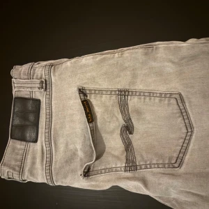 Nudie jeans - Snygga grå nudie jeans med klassisk femficksdesign och en svart läderdetalj vid midjan. 