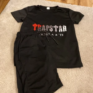 Svart t-shirt med tryck - Säljer en svart t-shirt och matchande shorts  från Trapstar i storlek S. Den är gjord av 100% polyester och har ett coolt tryck i rött och vitt med texten 'IT'S A SECRET'. Perfekt för en avslappnad stil eller streetwear-look. T-shirten är i bra skick och redo för nya äventyr!