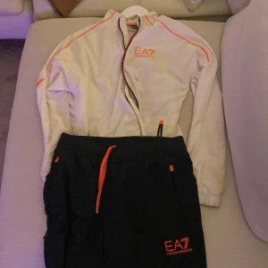 Svart och vit träningsdräkt från EA7 - Säljer en snygg träningsdräkt från EA7 Emporio Armani. Jackan är vit med orange detaljer och dragkedja, medan byxorna är svarta med matchande orange detaljer. Perfekt för träning eller en avslappnad dag. Både jackan och byxorna har EA7-loggan. Superbekväm och stilren!
