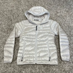 J.Lindeberg damjacka - Säljer en snygg vit pufferjacka från J.Lindeberg i storlek M. Jackan har en glansig finish och är perfekt för kyliga dagar. Den har en dragkedja framtill och en praktisk huva. Perfekt för både vardag och lite finare tillfällen. 🧥✨