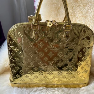 Guldig handväska med monogram MM - Säljer en superglammig guldig handväska från Louis Vuitton. Väskan har ett snyggt monogrammönster och är i mycket bra skick. Den har en dragkedja upptill och små handtag. Perfekt för att lyfta vilken outfit som helst! ✨