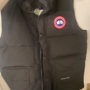 Svart dunväst från Canada Goose - Säljer en svart dunväst från Canada Goose nysick Västen har en dragkedja och knappar framtill samt två praktiska fickor. Perfekt för kyliga höst- och vinterdagar. Den har en klassisk stil med det ikoniska märket på bröstet. Passar både till vardags och mer äventyrliga aktiviteter.