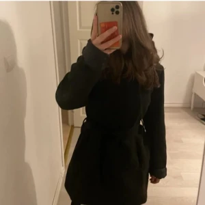 Svart kappa med huva - Säljer en stilren svart kappa med huva. Perfekt för höst och vinter, håller dig varm och snygg. Jackan har en normal passform och långa ärmar. Den är i mycket bra skick. Pris kan diskuteras