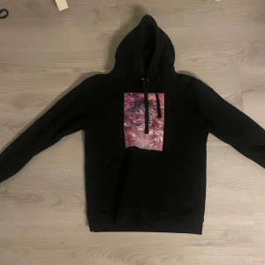 Svart hoodie med konstnärligt tryck - Säljer en svart hoodie med ett unikt konstnärligt tryck i rosa och lila på framsidan. Perfekt för att ge din outfit en cool och kreativ touch. Hoodien har en klassisk passform med långa ärmar och en bekväm huva. Perfekt för höst och vinter!